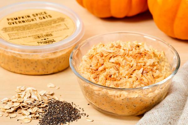 Glass bowl and plastic tub with label reading "Pumpkin Spice Overnight Oats" both filled with prepared oats, with dry oats, chia seeds, and pumpkins on the side