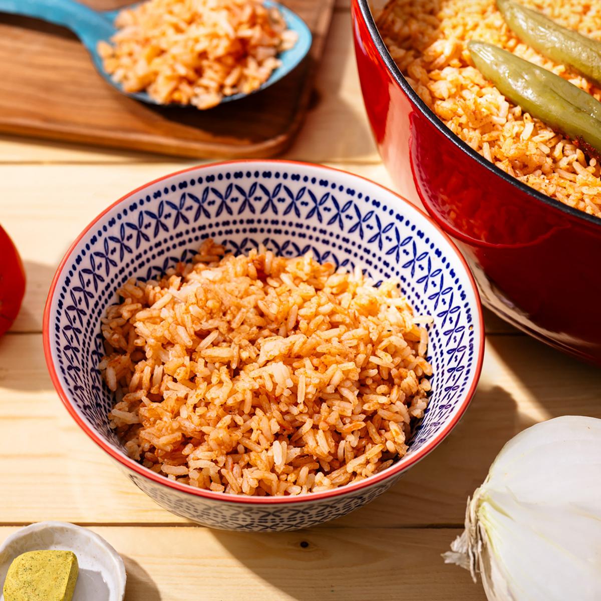 Bowl of Arroz Rojo (Mexican red rice) with fluffy tomato-seasoned rice, served on a wooden table with a pot of rice, onion, bouillon cube, and peppers in the background.