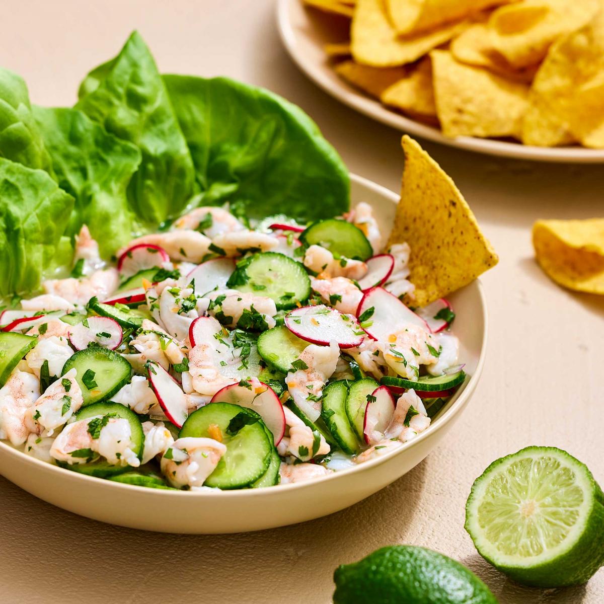 Shrimp ceviche in a bowl with lettuce, cucumbers, and a chip, with more chips and limes on the side