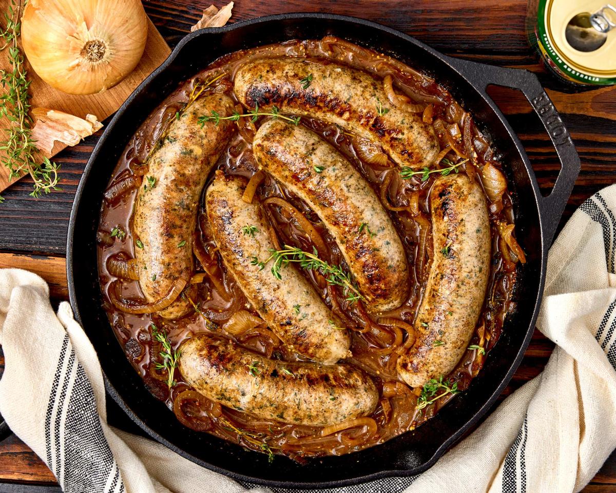 Fresh to Market beer and Bratwurst sausages braised in onion gravy with fresh thyme in a cast iron skillet, surrounded by onions and herbs on a wood table