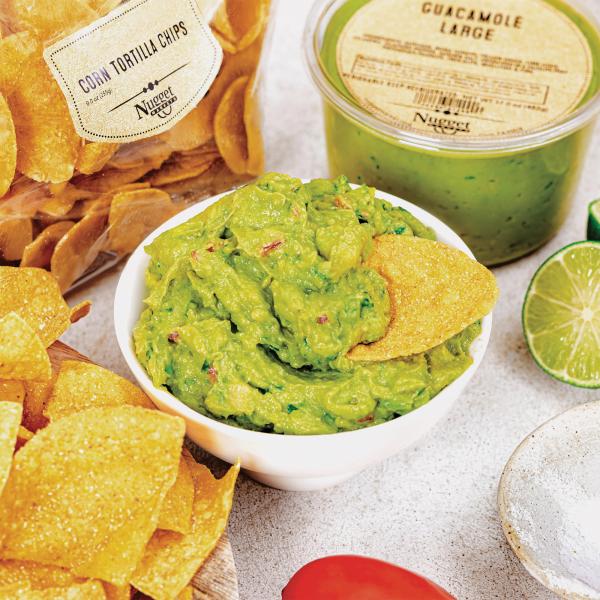 Bowl of guacamole with a chip dipping in it surrounded by chips, a bag of Fresh to Market Tortilla Chips, a tube of Fresh to Market Guacamole, a lime, a tomato, and salt