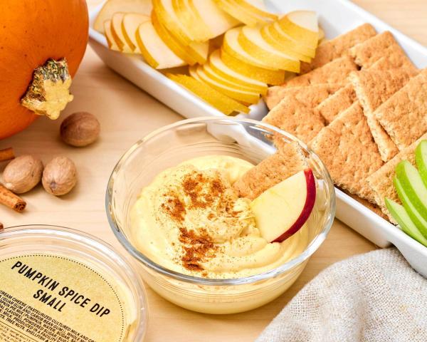Glass bowl of dip with apple slice and graham cracker, plus plastic tub with label reading "Pumpkin Spice Dip Small" and pumpkin, cinnamon sticks, nutmeg, and tray of graham crackers and apple slices on the side