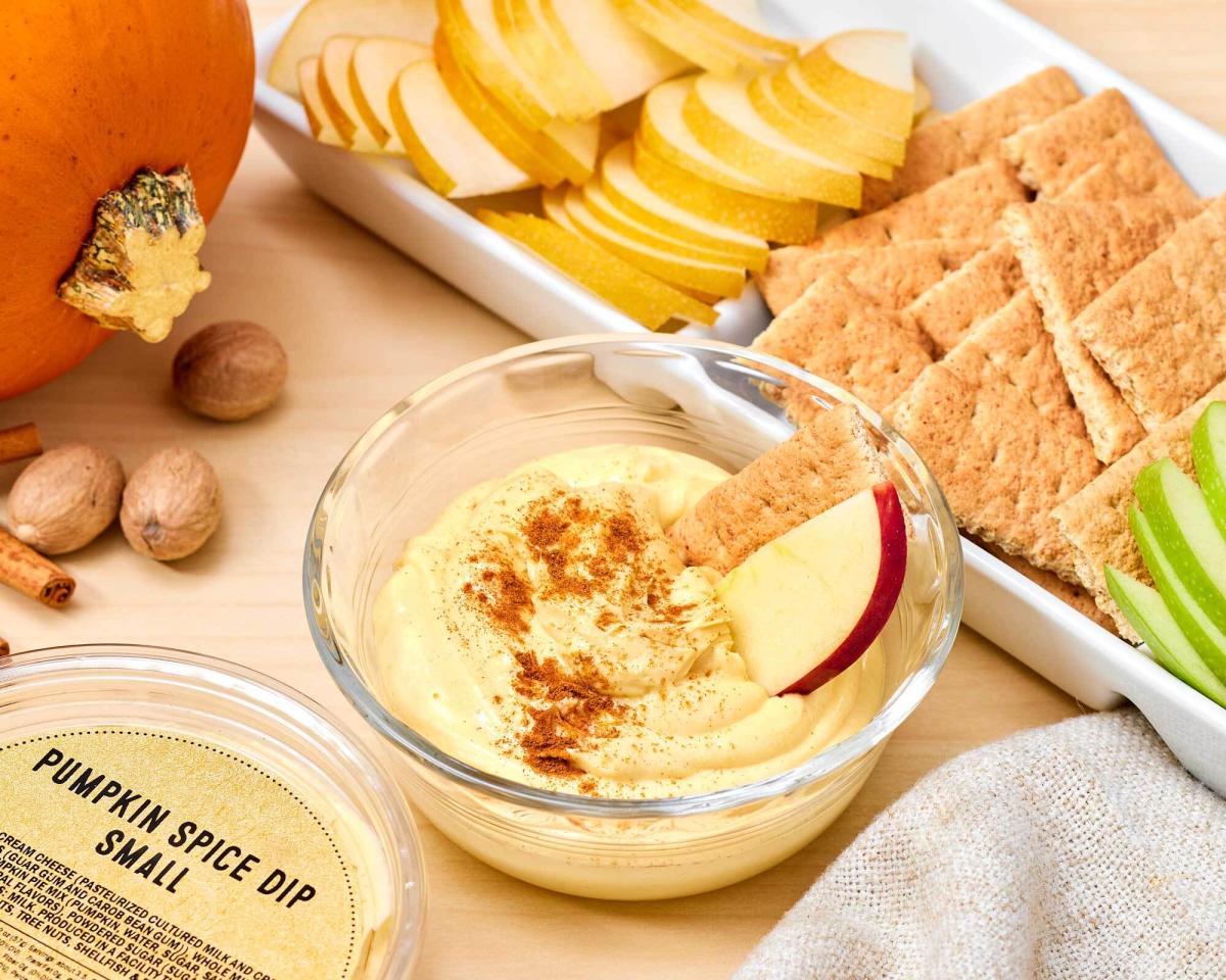 Glass bowl of dip with apple slice and graham cracker, plus plastic tub with label reading "Pumpkin Spice Dip Small" and pumpkin, cinnamon sticks, nutmeg, and tray of graham crackers and apple slices on the side