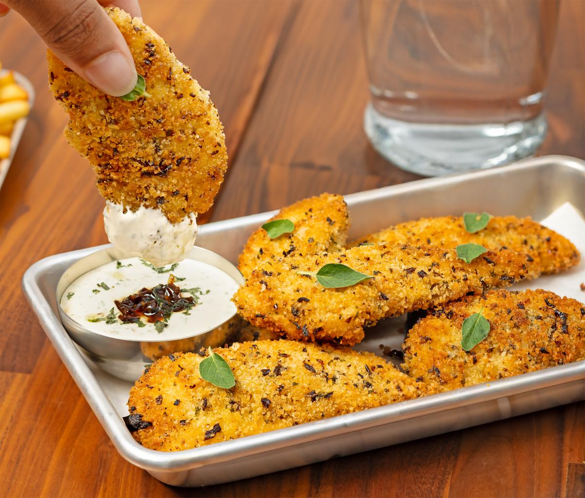 Hand dipping a battered chicken tender into creamy dip on a metal tray holding more chicken tenders
