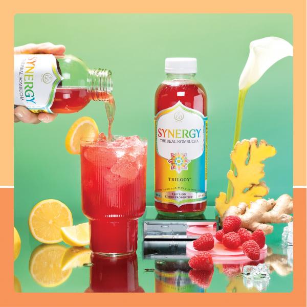 Hand pouring bottle of GT's Kombucha into glass with another bottle, lemons, raspberries and ginger around it