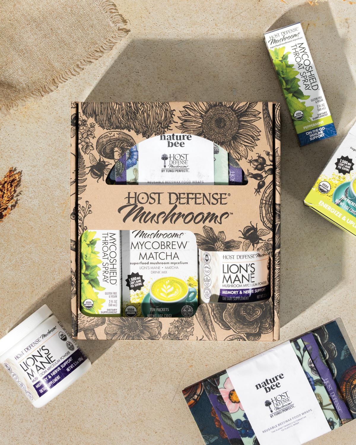 Flat-lay product photo of a Host Defense Mushrooms gift box featuring MycoShield Throat Spray, MycoBrew Matcha drink mix, and Lion's Mane mycelium powder, surrounded by additional product boxes and Nature Bee reusable beeswax food wraps, styled on a 