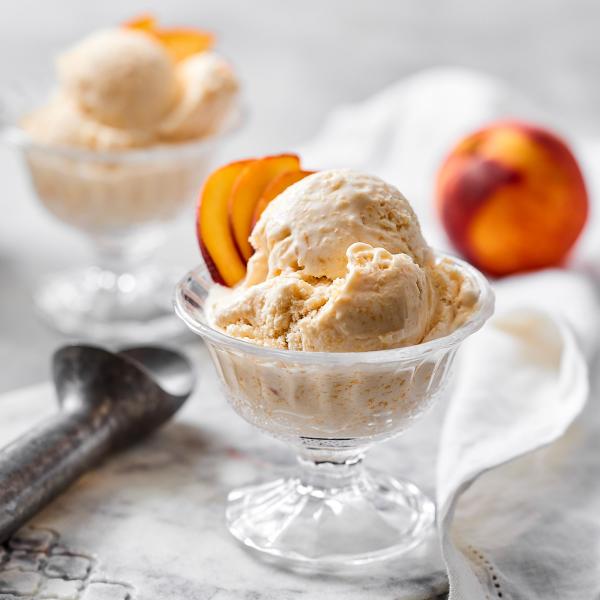 Two glass dessert cups of peach ice cream topped with fresh peach slices, with a whole peach and ice cream scoop in the background