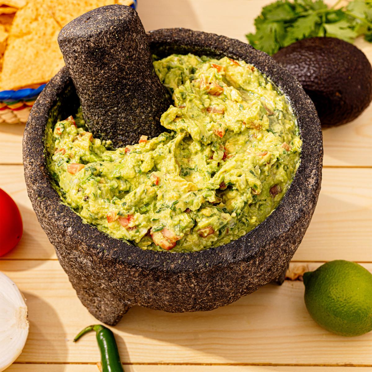 Guacamole Clasico served in a traditional black stone molcajete with a matching pestle, surrounded by fresh avocados, lime, cilantro, tortilla chips, tomato, onion, and a jalape&ntilde;o on a light wood surface.