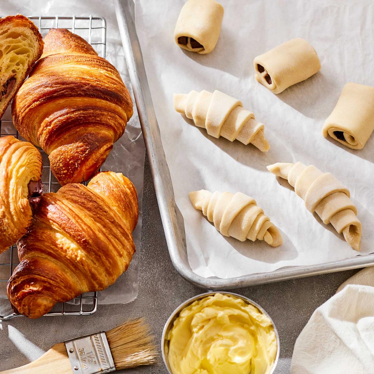 Yeast Laminated dough shaped into croissants on a baking sheet with baked croissants on side and brush wih buter