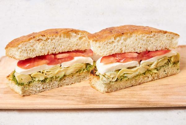 Milano vegetarian sandwich cut in half on a wooden board, showing layers of pesto, marinated artichoke hearts, fresh mozzarella, and roasted tomatoes on focaccia.