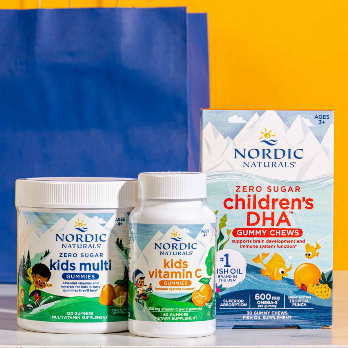 Nordic Naturals Zero Sugar Kids Multi Gummies, Kids Vitamin C Gummies, and Zero Sugar Children's DHA Gummy Chews in front of blue paper bag and orange background