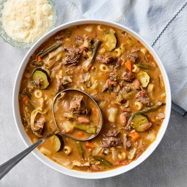 Overhead view of a hearty minestrone soup in a white bowl, filled with braised beef, zucchini, green beans, carrots, borlotti beans, cabbage, and ditalini pasta in a rich tomato broth. A ladle rests in the bowl; a small dish of grated Parmesan sits alongs