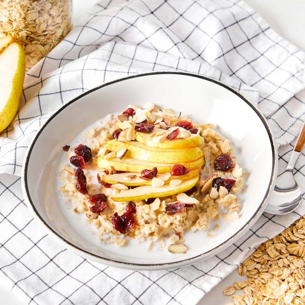 Bowl of oatmeal topped with sliced pear and dried cranberries, with jar of oats, pear half, cloth napkin and spoon on the side
