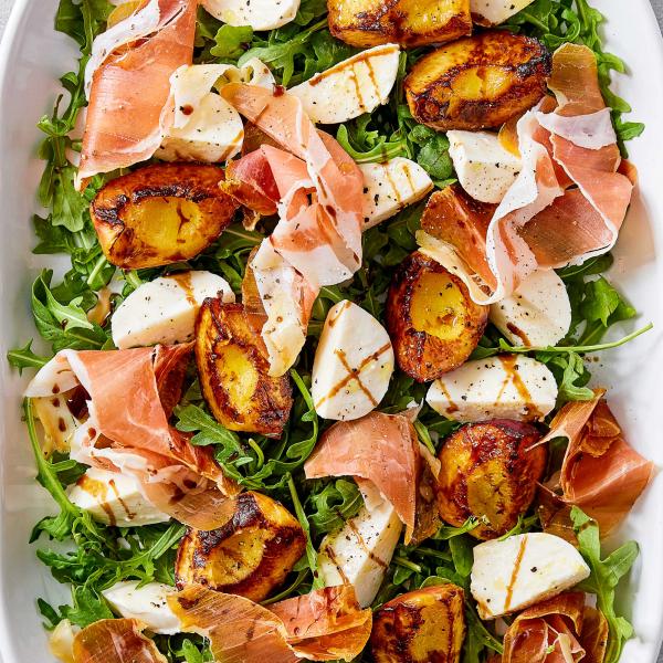 Platter of grilled peach halves, fresh mozzarella, and prosciutto on a bed of arugula drizzled with balsamic glaze