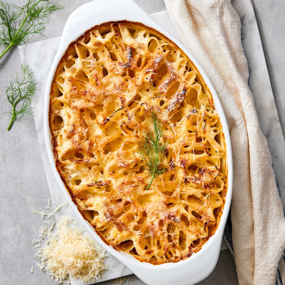 Golden-brown potato and fennel gratin in a white oval baking dish, with a lacy caramelized cheese crust, garnished with a fresh fennel frond. Shredded cheese and fennel sprigs alongside on a gray surface.