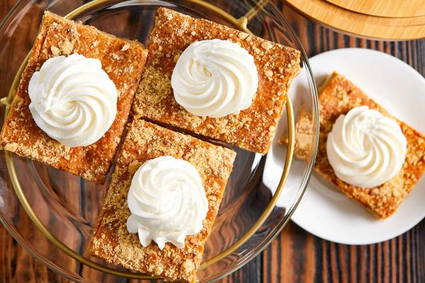 Bird's eye view of three pumpkin bars on a glass plate stand over a fourth bar on a white plate. Each pumpkin bar is topped with a swirl of whipped cream.