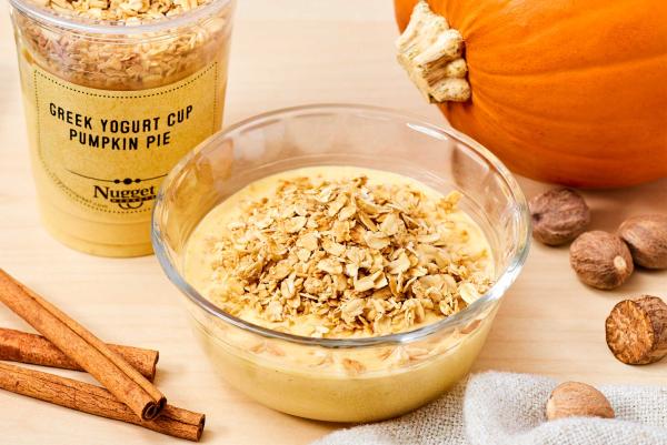 Glass bowl and plastic cup with label reading "Greek Yogurt Cup Pumpkin Pie", both filled with yogurt and granola, with cinnamon sticks and a pumpkin on the side