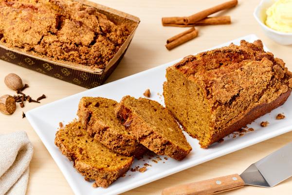 Whole pumpkin spice loaf cake in a paper loaf form, plus a partially sliced loaf cake on a plate next to whole spices, a metal spatula, and a small bowl of butter