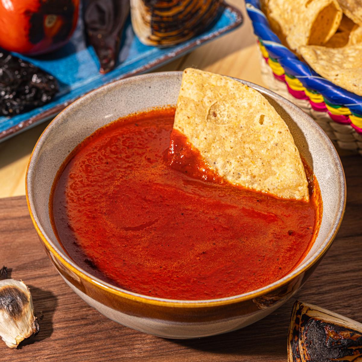 Salsa Rojo served in a ceramic bowl with a tortilla chip for dipping, surrounded by tortilla chips, roasted vegetables, garlic, and grilled peppers on a rustic wooden table.