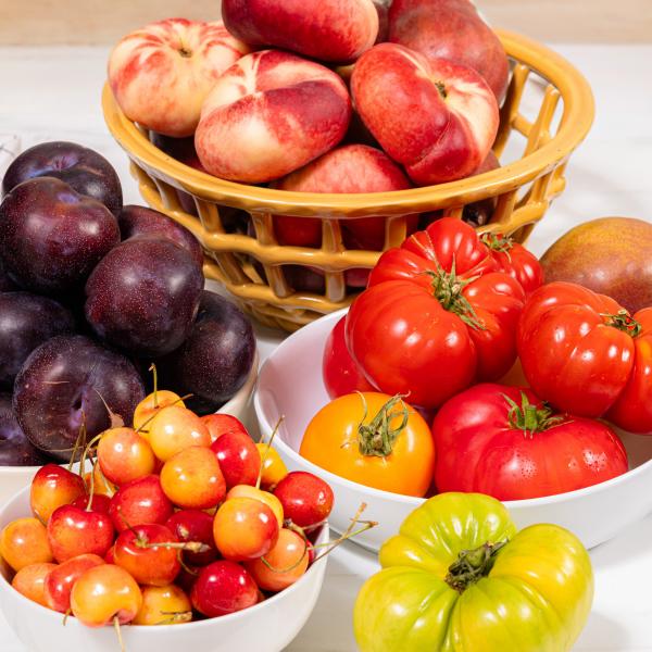 Bowls of fresh summer produce including heirloom tomatoes, cherries, plums, and donut peaches
