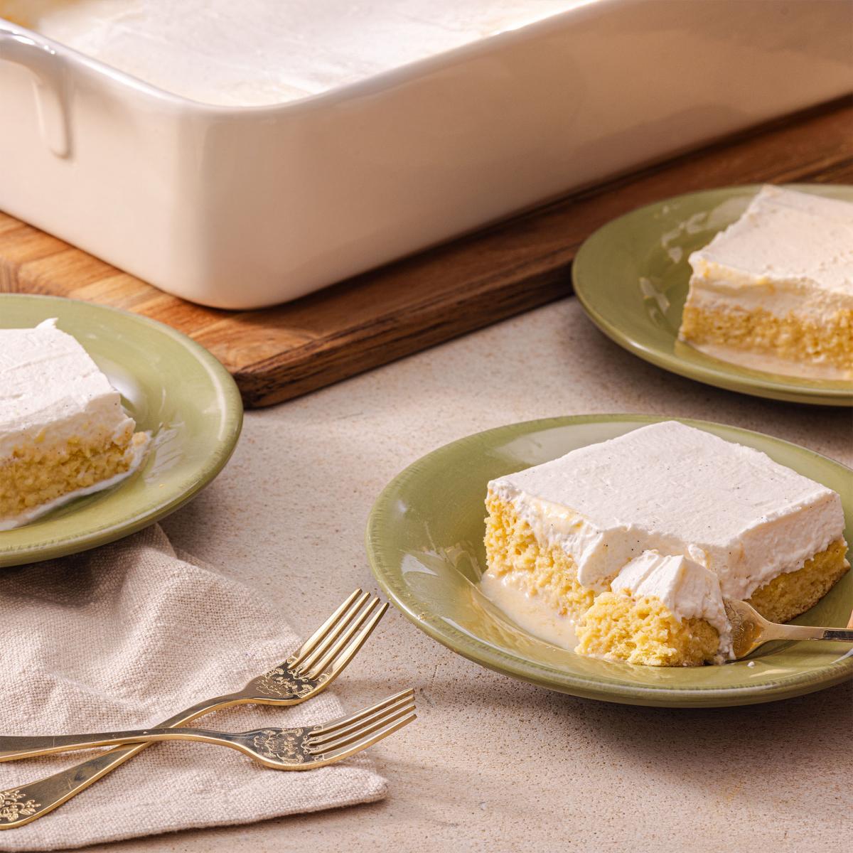 Slice of tres leches cake with whipped cream topping on a green plate, gold fork alongside.