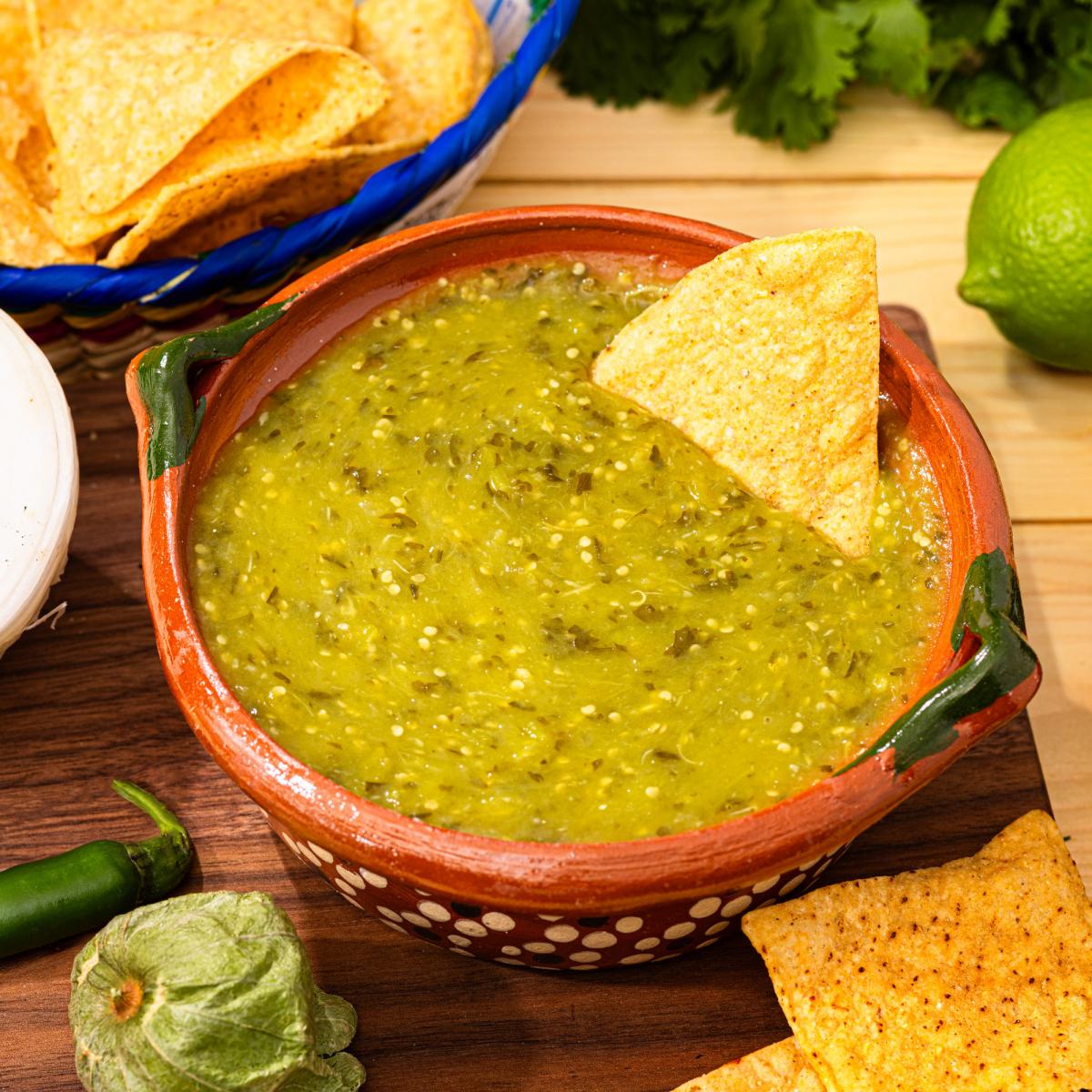 Salsa Verde served in a traditional ceramic bowl with a tortilla chip for dipping, surrounded by tortilla chips, tomatillos, jalape&ntilde;o, cilantro, and lime on a rustic wooden table.
